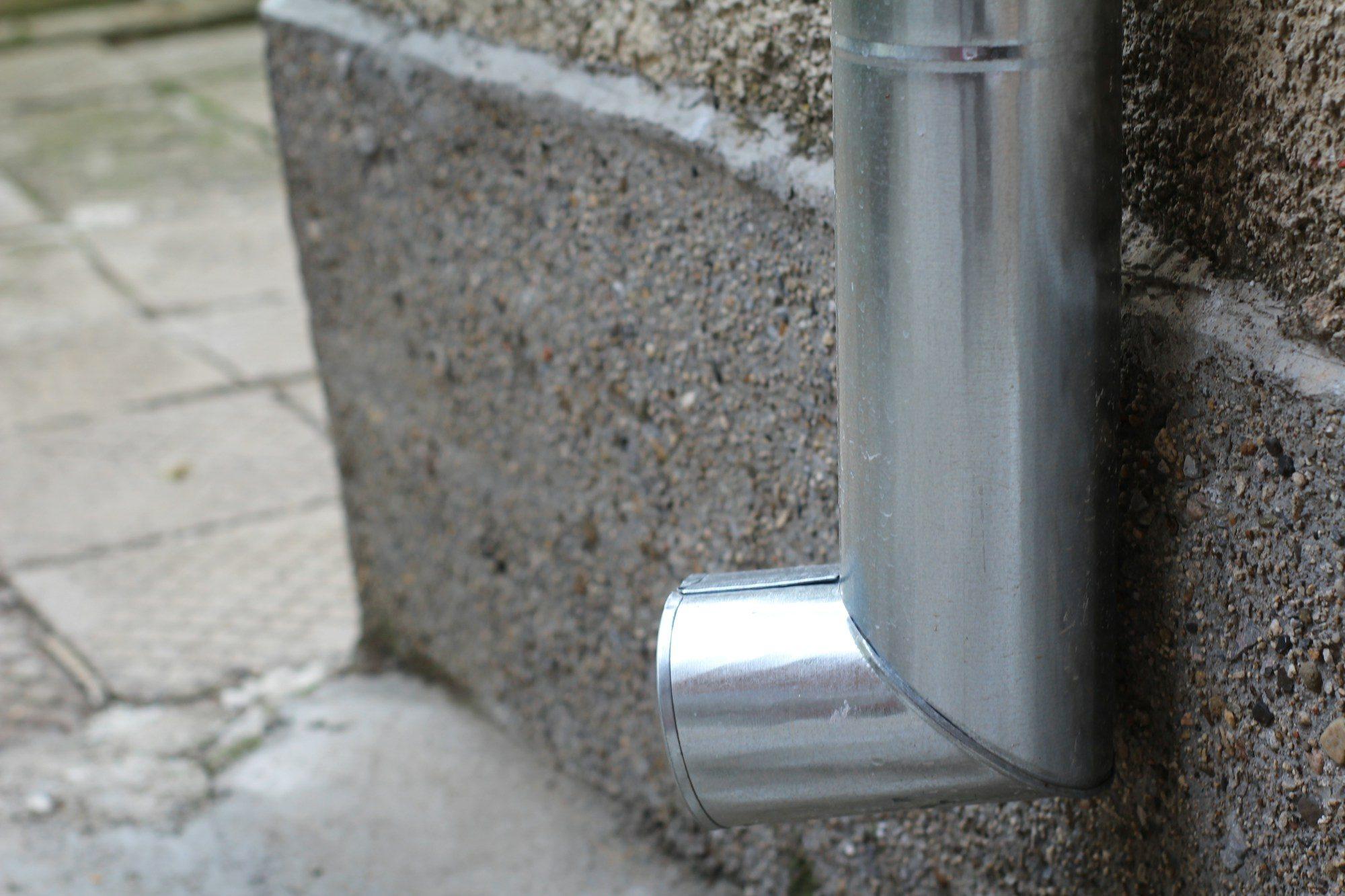 Metal downspout elbow at the base of a concrete wall discharging onto paving stones